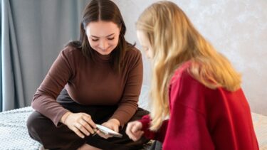 To unge piger sidder på en seng og kigger på en smartphone