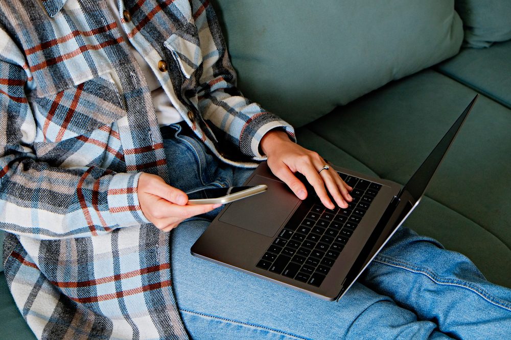 Kvinde i sofa med laptop på skødet tjekker mobilen