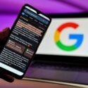 En hånd holder en smartphone, i baggrunden ses en laptop med et google-logo
