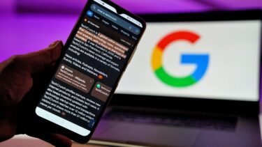 En hånd holder en smartphone, i baggrunden ses en laptop med et google-logo