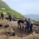 En gruppe arkæologer og arkæologistuderende graver udgravningssted ved kysten på græsklædte bakker med værktøj og spande spredt rundt omkring. Havet og et hus på en bjergskråning ses i baggrunden under en overskyet himmel.