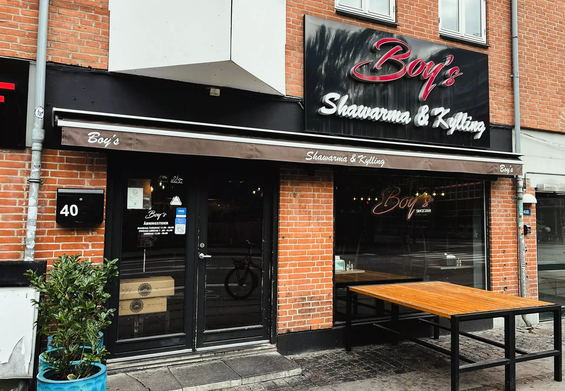 En murstensbutiksfacade med en sort dør og vinduer, en brun markise med teksten 'Boys Shawarma & Kylling' og skilte med teksten 'Boy's'. En blå potteplante og et udendørs træbord er placeret nær indgangen.