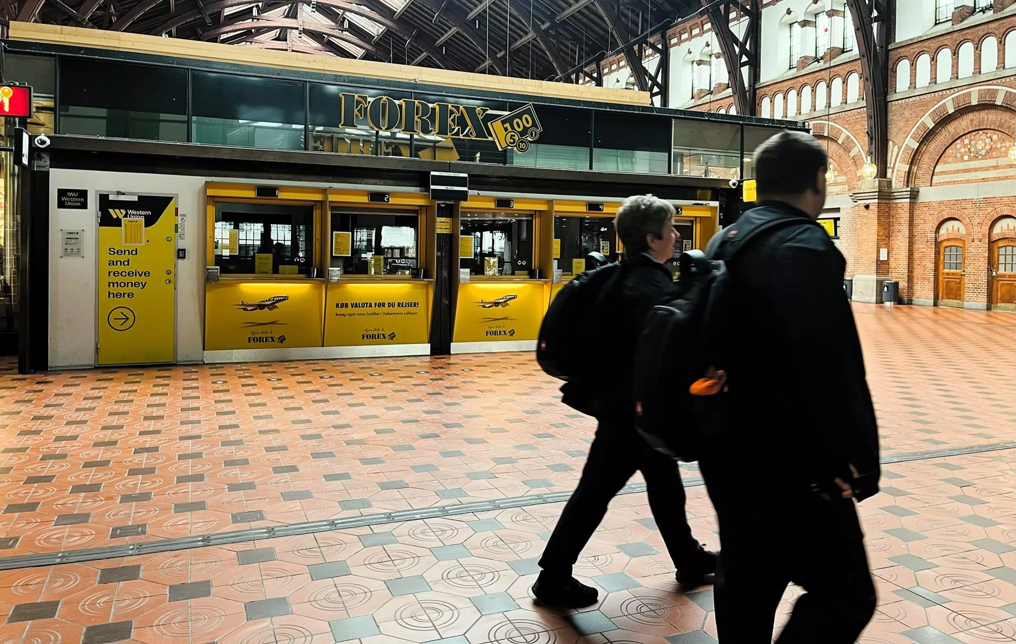 To personer med rygsække går forbi en gul valutavekslingsboks inde i en rummelig indendørs togstation med mønstret flisebelagt gulv og høje, buede vinduer.