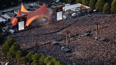 Luftfoto af et stort udendørs koncertpublikum foran en stor orange teltscene, Orange Scene på Roskilde Festival. Omgivet af træer og eventstrukturer på en solrig dag.