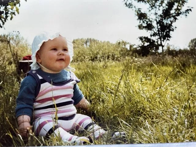 En baby iført hvid kyse og farverigt stribet tøj sidder i højt græs udendørs og smiler. Træer og grønne områder ses i baggrunden på en solrig dag.