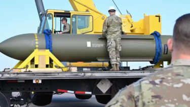 Militærpersonale i camouflageuniformer overvåger en stor bombe, der er sikret på en lastbil, med en gul kran i baggrunden på en klar dag.