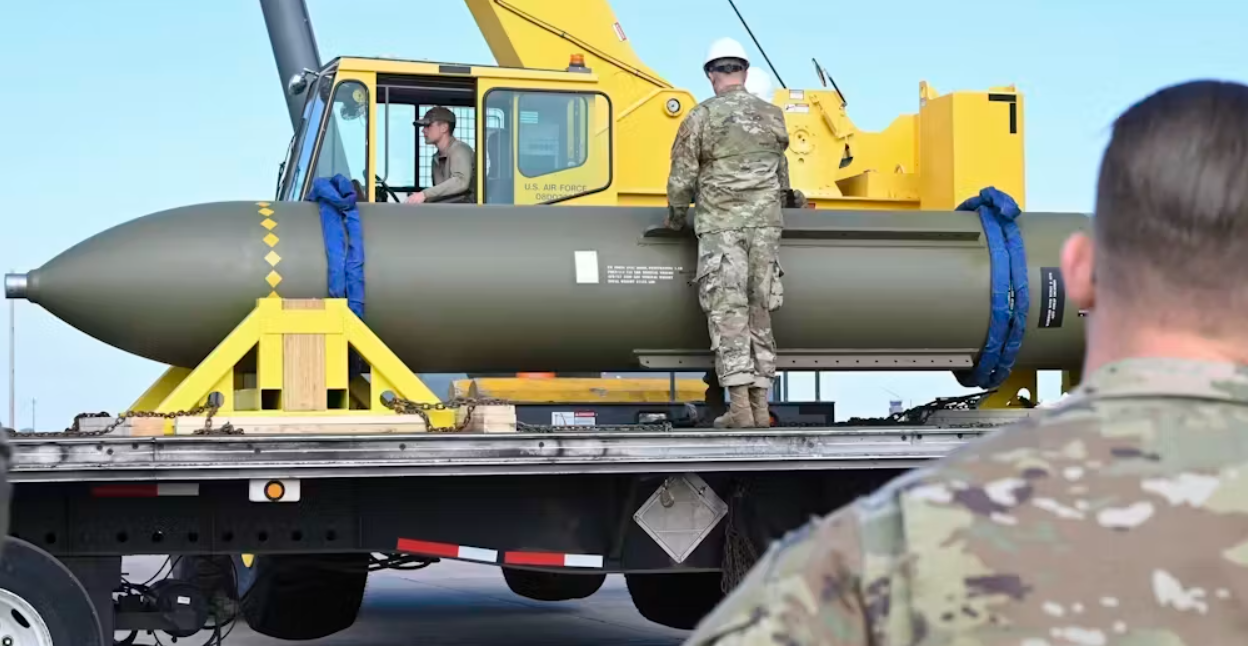 Militærpersonale i camouflageuniformer overvåger en stor bombe, der er sikret på en lastbil, med en gul kran i baggrunden på en klar dag.