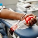 En person donerer blod ved at klemme på en rød stressbold, mens blodet løber gennem et rør ned i en opsamlingspose. Medicinsk udstyr er synligt i baggrunden.