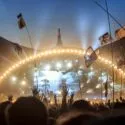 Publikum på Roskilde Festival løfter hænderne og vifter med flag foran en stærkt oplyst scene med en stor buet struktur mod en blå aftenhimmel.