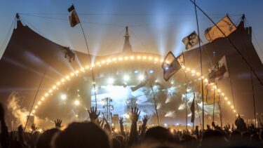 Publikum på Roskilde Festival løfter hænderne og vifter med flag foran en stærkt oplyst scene med en stor buet struktur mod en blå aftenhimmel.