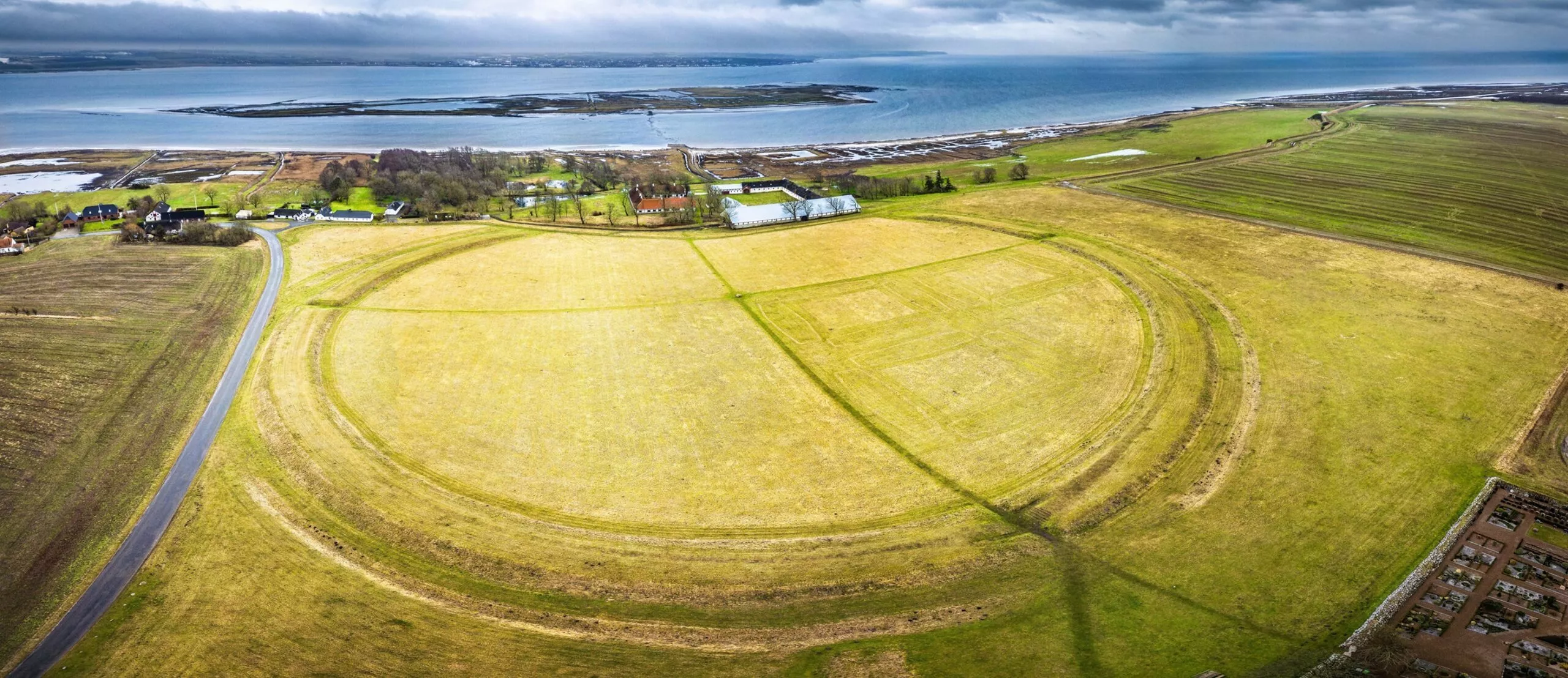 Luftfoto af et stort cirkulært omrids på en græsmark nær kysten med et par bygninger, en vej og vand i baggrunden under en overskyet himmel.