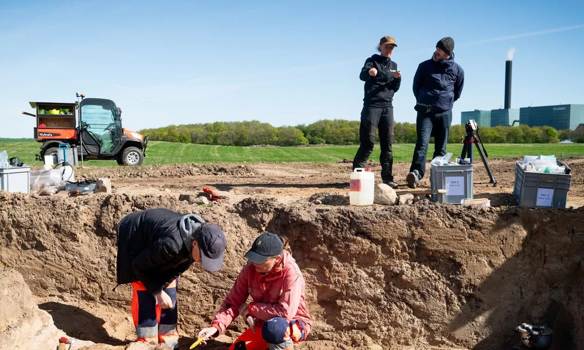 To arkæologer arbejder på en jordudgravning, mens to personer står i nærheden og taler sammen. Udstyr, kasser og et erhvervskøretøj er synlige, med grønne marker og en industribygning i baggrunden under en klar blå himmel.