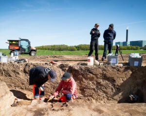 To arkæologer arbejder på en jordudgravning, mens to personer står i nærheden og taler sammen. Udstyr, kasser og et erhvervskøretøj er synlige, med grønne marker og en industribygning i baggrunden under en klar blå himmel.