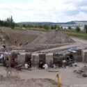 En gruppe arkæologer udgraver et stort åbent område med dybe grøfter og jordsøjler, omgivet af træer, bakker og en vandmasse under en delvis overskyet himmel. Udstyr og redskaber ligger spredt rundt omkring i området.