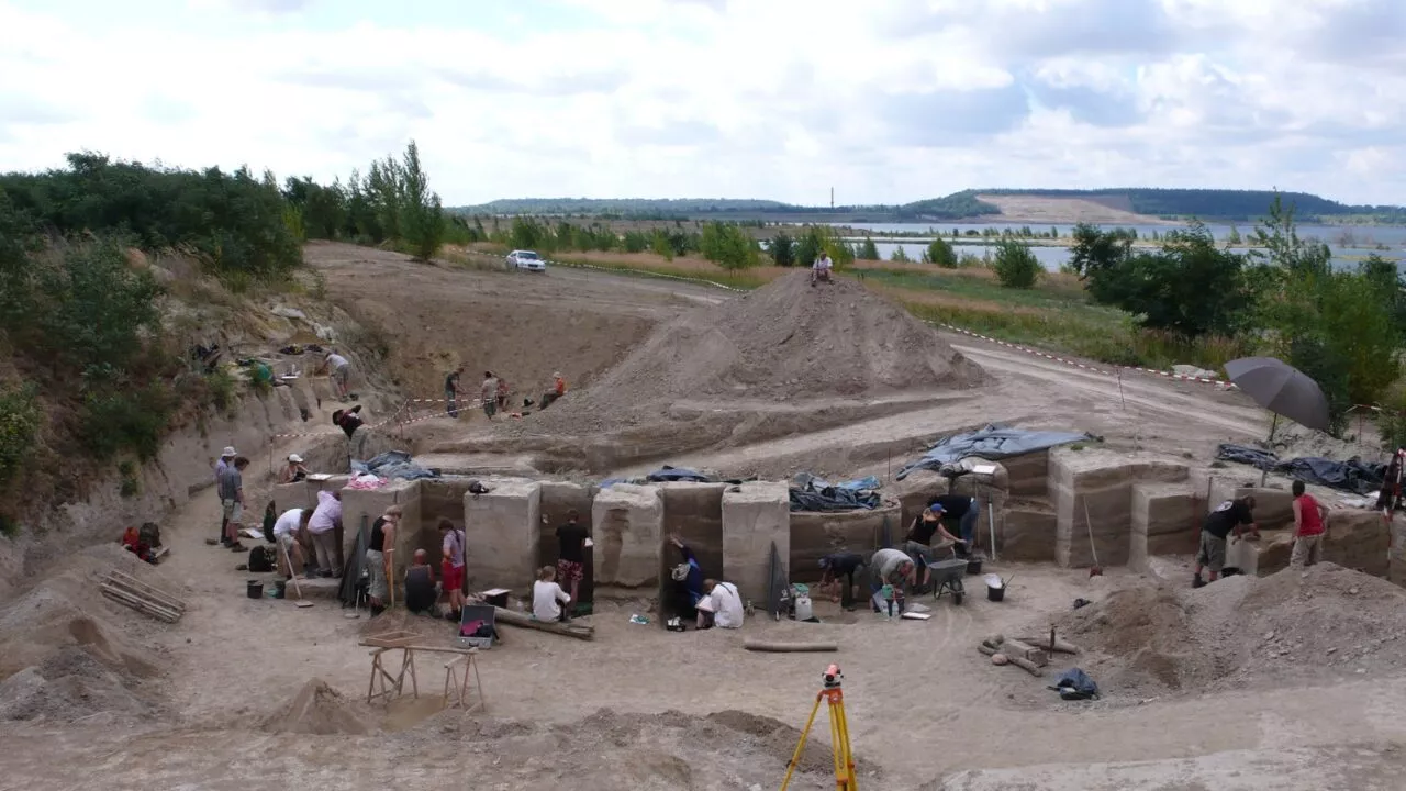 En gruppe arkæologer udgraver et stort åbent område med dybe grøfter og jordsøjler, omgivet af træer, bakker og en vandmasse under en delvis overskyet himmel. Udstyr og redskaber ligger spredt rundt omkring i området.