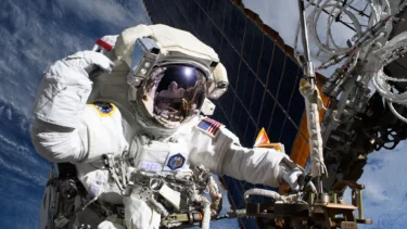 Astronaut Anne McClain i en hvid rumdragt svæver i rummet og arbejder på udstyr uden for et rumfartøj med et stort solpanel i baggrunden. Jorden og skyerne er synlige nedenfor. Astronautens visir reflekterer en anden person.