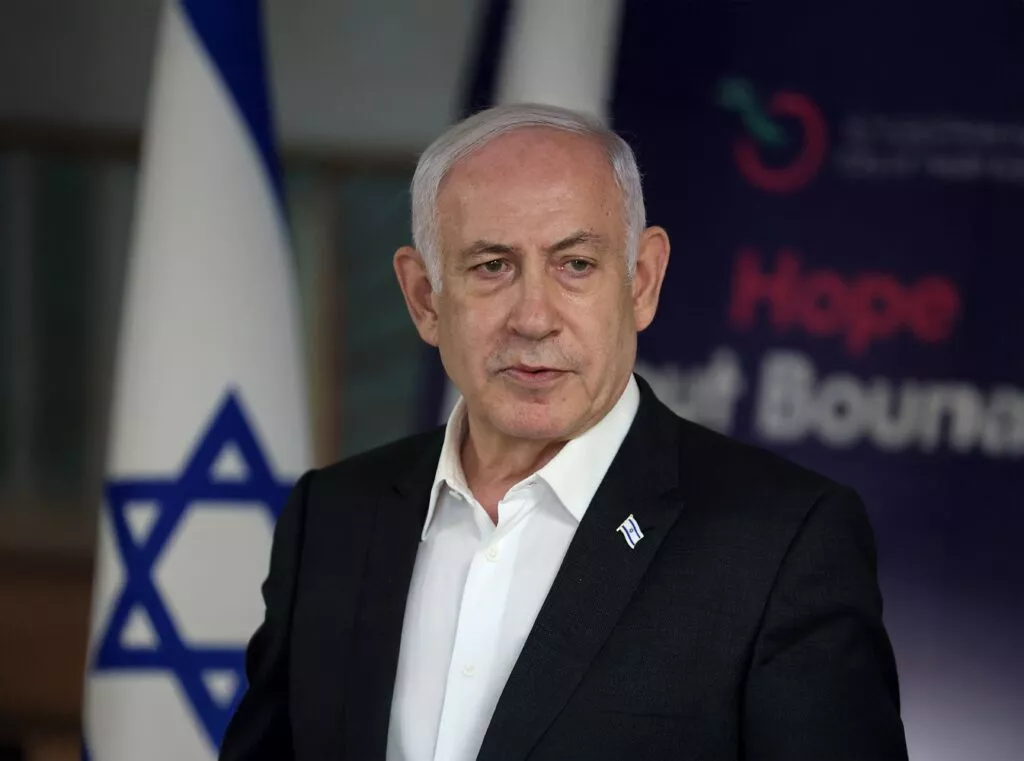 Benjamin Netanyahu i sort jakkesæt og hvid skjorte står foran et israelsk flag og et skilt med ordet Hope i baggrunden.