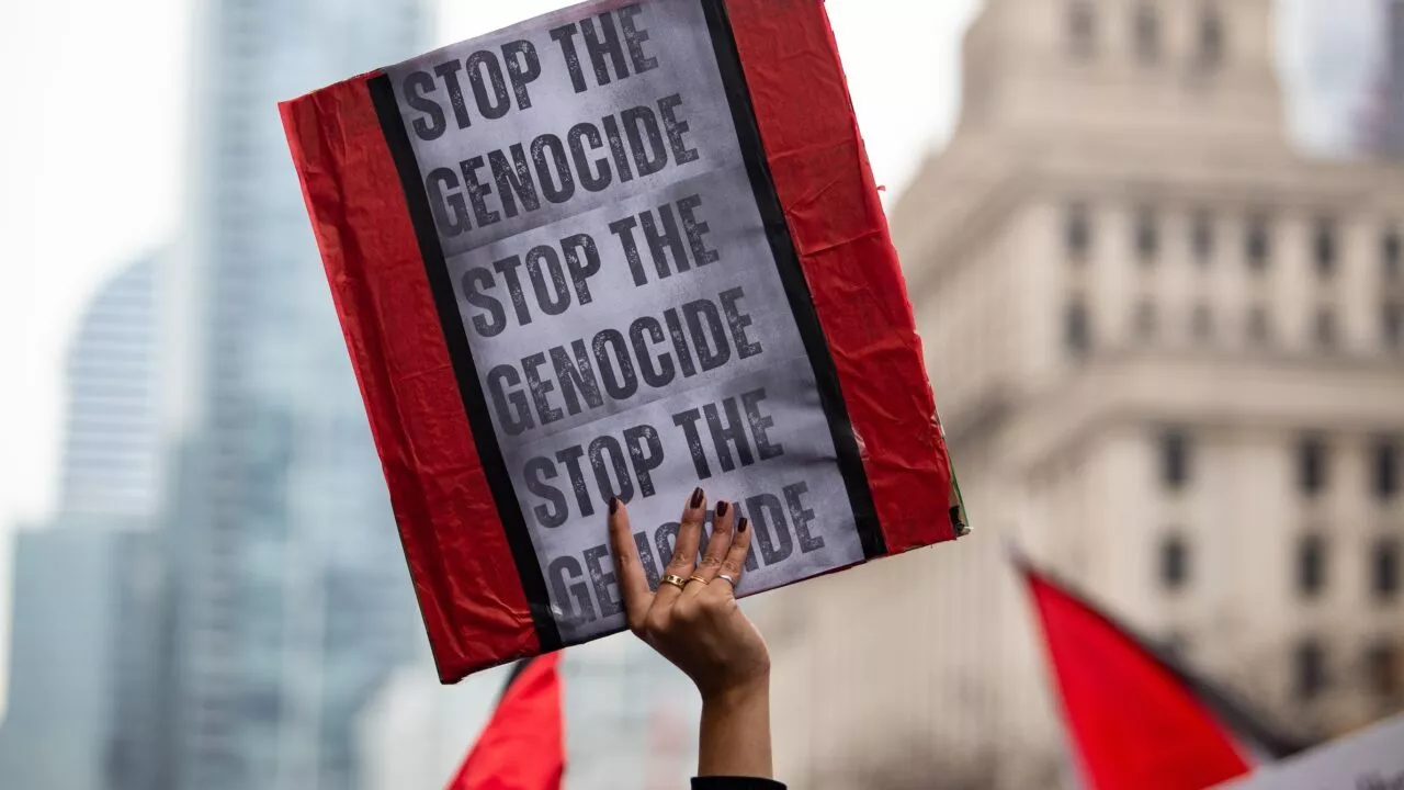 En person holder et skilt med teksten STOP THE GENOCIDE op flere gange under en demonstration med røde kanter på skiltet og slørede bybygninger i baggrunden.