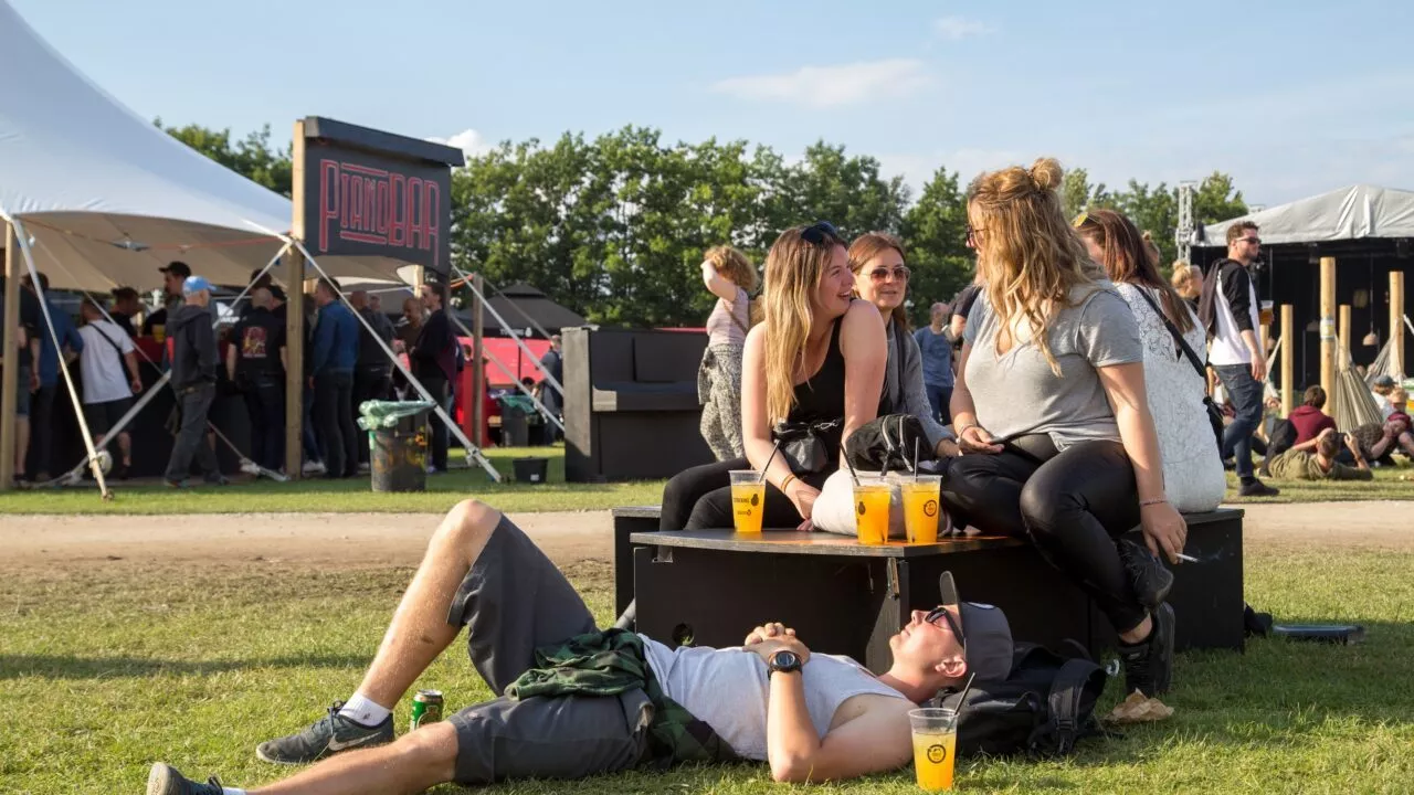 En gruppe unge mennesker sidder og snakker på en bænk på en udendørs festival, med drinks på bordet og en person liggende på græsset, mens en folkemængde samles nær telte og boder i baggrunden på en solrig dag.