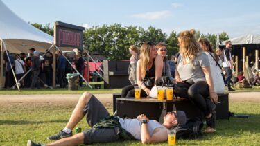 En gruppe unge mennesker sidder og snakker på en bænk på en udendørs festival, med drinks på bordet og en person liggende på græsset, mens en folkemængde samles nær telte og boder i baggrunden på en solrig dag.