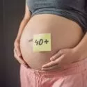 En gravid person iført grå skjorte og lyserøde bukser holder om sin mave, som har en gul seddel med påskriften 40+, hvilket tyder på graviditet i alderen 40 år eller derover.