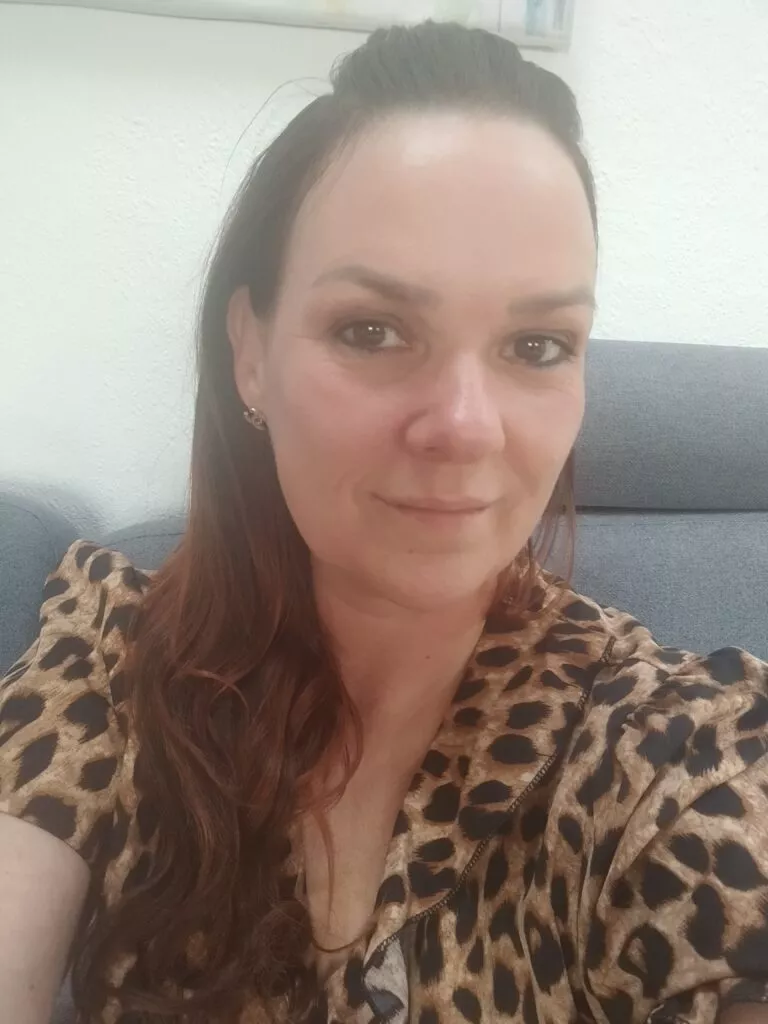 En kvinde med langt brunt hår og lys hudfarve sidder i en grå sofa og smiler let. Hun er iført en top med leopardprint og små øreringe, og i baggrunden ses en lys væg.
