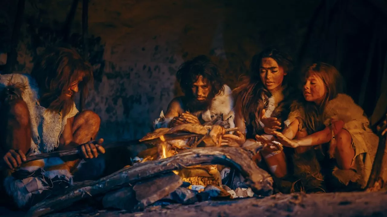 Fire personer klædt som forhistoriske mennesker sidder omkring et bål inde i en hule, hvor de varmer deres hænder og rister mad, mens den dæmpede belysning fremhæver deres pelstøj og primitive omgivelser.