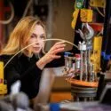 En kvinde med blondt hår arbejder intenst med maskiner og udstyr i et laboratorium eller i industrien, mens hun justerer knapper og ledninger, omgivet af tekniske instrumenter.