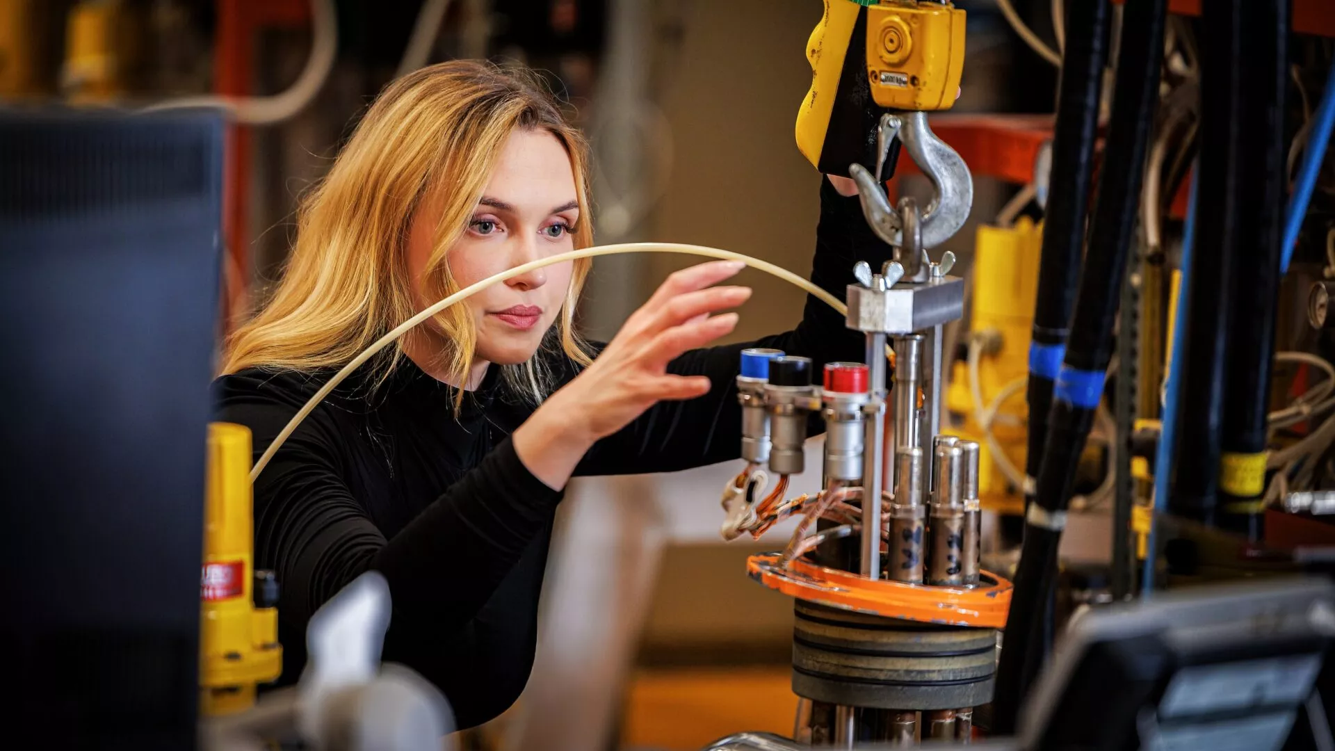 En kvinde med blondt hår arbejder intenst med maskiner og udstyr i et laboratorium eller i industrien, mens hun justerer knapper og ledninger, omgivet af tekniske instrumenter.