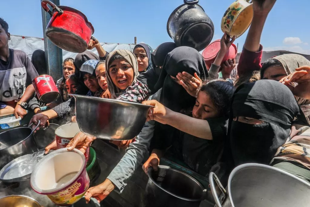 En gruppe kvinder og børn i Gaza holder tomme gryder og skåle frem i et overfyldt udendørs miljø under en lys himmel, hvilket tyder på, at de venter på at få mad udleveret.