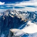 Snedækkede bjergtoppe ved Mont Blanc under en lyseblå himmel med spredte skyer, forrevne klipperygge og fjerne bjergkæder i baggrunden. Scenen giver en følelse af storhed og naturlig skønhed.