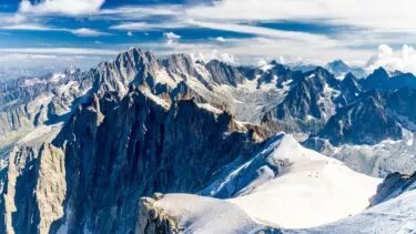 Snedækkede bjergtoppe ved Mont Blanc under en lyseblå himmel med spredte skyer, forrevne klipperygge og fjerne bjergkæder i baggrunden. Scenen giver en følelse af storhed og naturlig skønhed.