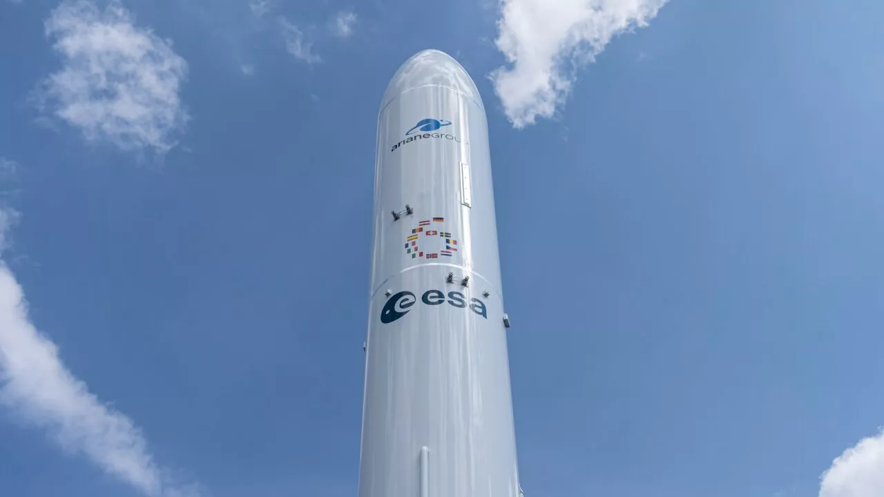 Et nærbillede af en hvid raket mod en blå himmel med skyer, med logoer for Arianespace og ESA (European Space Agency) på kroppen.