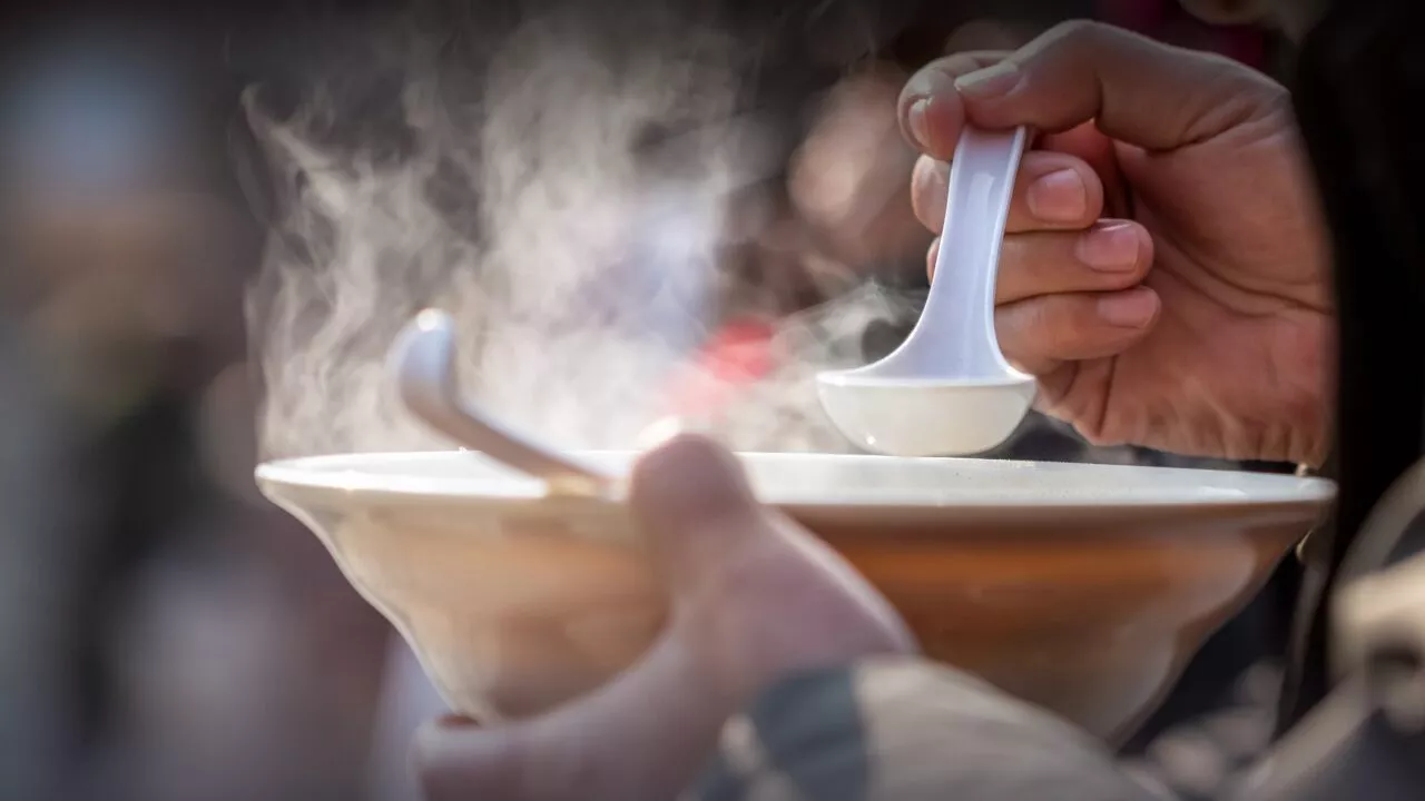 En person holder en dampende skål i den ene hånd og bruger en hvid ske med den anden hånd, klar til at spise varm suppe. Scenen er et nærbillede med synlig opstigende damp.