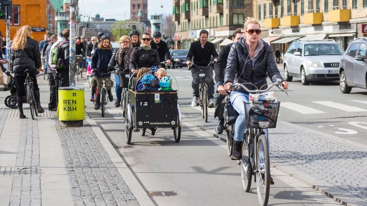 Folk cykler på en travl cykelsti i byen, bl.a. en kvinde med solbriller og en ladcykel med børn, med fodgængere og bybygninger i baggrunden.