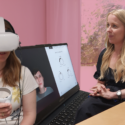 En kvinde med et VR-headset holder en controller, mens en anden kvinde med langt blondt hår sidder ved siden af en bærbar computer, der viser en virtuel avatar og kontrolindstillinger, begge i et lyserødt rum.