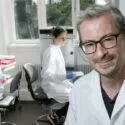 Tre forskere i hvide kitler arbejder i et lyst laboratorium. En mand med briller smiler til kameraet i forgrunden, mens de to andre arbejder med laboratorieudstyr i baggrunden. Sollyset strømmer ind gennem vinduerne.