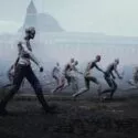 En gruppe zombielignende figurer med forrådnet, bleg hud går og vakler gennem et tåget, øde bylandskab med en stor, sløret bygning i baggrunden.