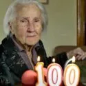 En ældre kvinde med hvidt hår sidder ved siden af en rollator bag en kage med tændte lys formet som tallet 100 og fejrer sin 100-års fødselsdag i hjemmet.