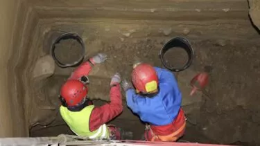 To arkæologer iført hjelme og lyst sikkerhedsudstyr graver forsigtigt jord op i en dyb, rektangulær grube. Spande til jord og værktøj står i nærheden, og begge arbejder med handsker på den blottede jordvæg.