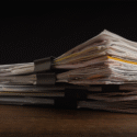 En stak uorganiserede papirer og dokumenter, der holdes sammen af store sorte clips, ligger på en træplade mod en mørk baggrund.