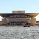 Billedet viser Københavns moderne operahus med det brede tag og glasfacaden set fra den anden side af vandet på en klar, solrig dag. Både er synlige på vandet foran bygningen.