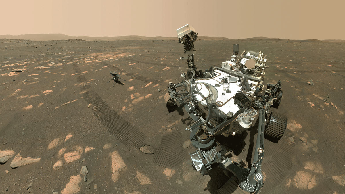 NASA's Perseverance-rover sidder på Mars' stenede, rødlige overflade med sin robotarm udstrakt. Ingenuity-helikopteren er i nærheden, og hjulsporene er synlige i Mars-jorden.