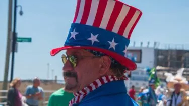 En mand klædt ud som Uncle Sam, iført en rød, hvid og blå høj hat med stjerner, solbriller og en blå jakke, står udendørs ved en parade med mennesker og en båd i baggrunden.