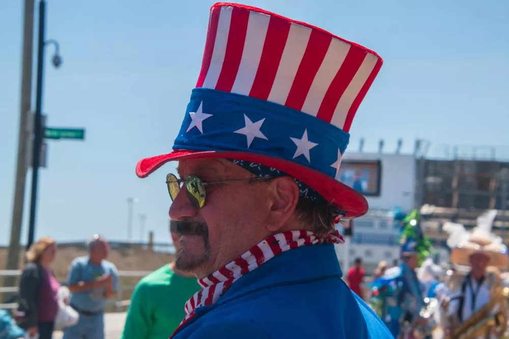 En mand klædt ud som Uncle Sam, iført en rød, hvid og blå høj hat med stjerner, solbriller og en blå jakke, står udendørs ved en parade med mennesker og en båd i baggrunden.