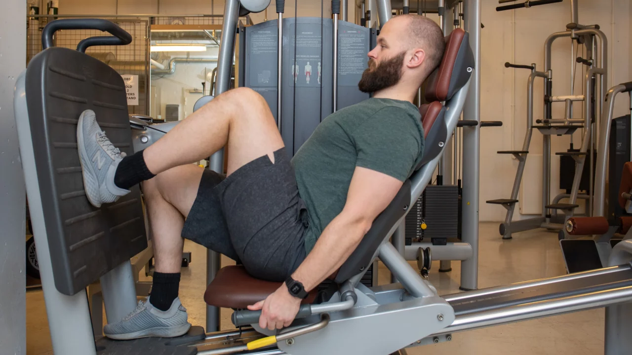 En mand med skæg og kort hår bruger en benpresmaskine i et fitnesscenter. Han sidder ned og er iført en grøn t-shirt, mørke shorts, grå sneakers og en aktivitetstracker. Træningsudstyret er synligt i baggrunden.