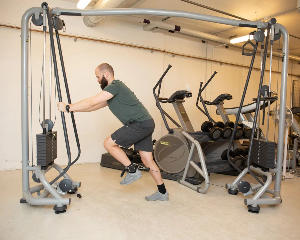 En mand med skæg træner på en kabelmaskine i et fitnesscenter, hvor han trækker i et håndtag med venstre hånd, mens han balancerer på et ben. Elliptiske maskiner er synlige i baggrunden.