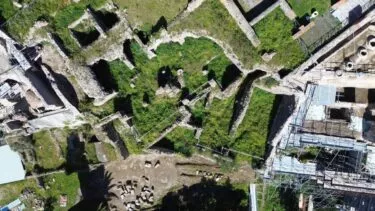 Luftfoto af gamle stenruiner, der er delvist overgroet med græs, med rektangulære og uregelmæssige stenfundamenter sammen med stilladser og udgravningsmaterialer.