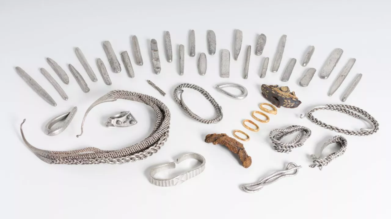 En samling af sølvbarrer, armbånd, ringe og andre smykker fra vikingetiden udstillet på en hvid overflade, hvor nogle af genstandene har indviklede mønstre, og der er en rusten metalgenstand blandt dem.