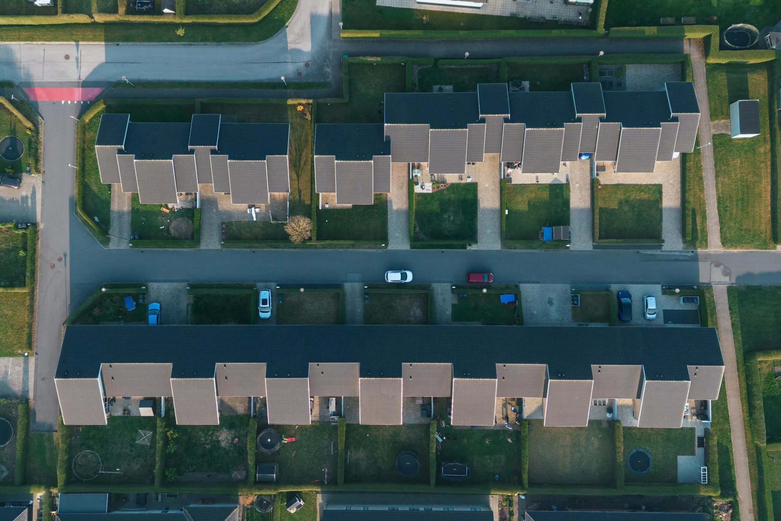 Luftfoto af et boligkvarter med rækker af identiske huse, indkørsler med parkerede biler og velholdte grønne plæner adskilt af hække.